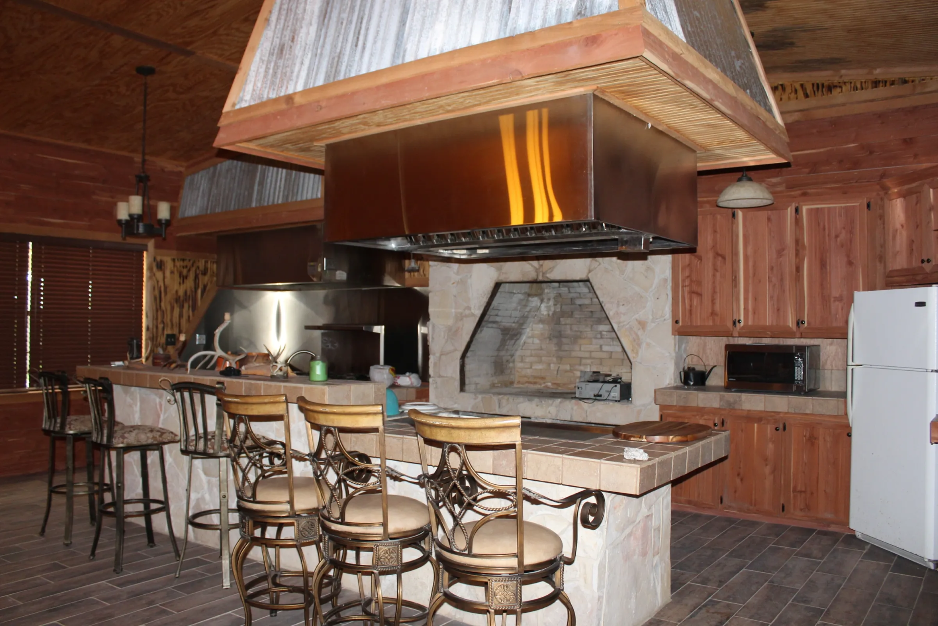 Rustic kitchen with a large island featuring tile countertop, ornate bar stools, wooden cabinets, a white refrigerator, a microwave, and a stone fireplace with a metal hood above the cooking area.