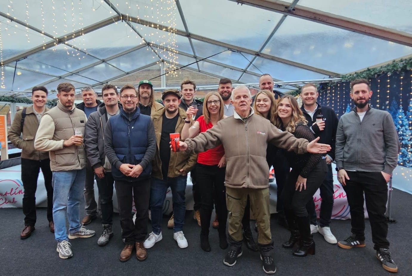 Group of happy people posing together under a tent decorated with string lights, some holding drinks.