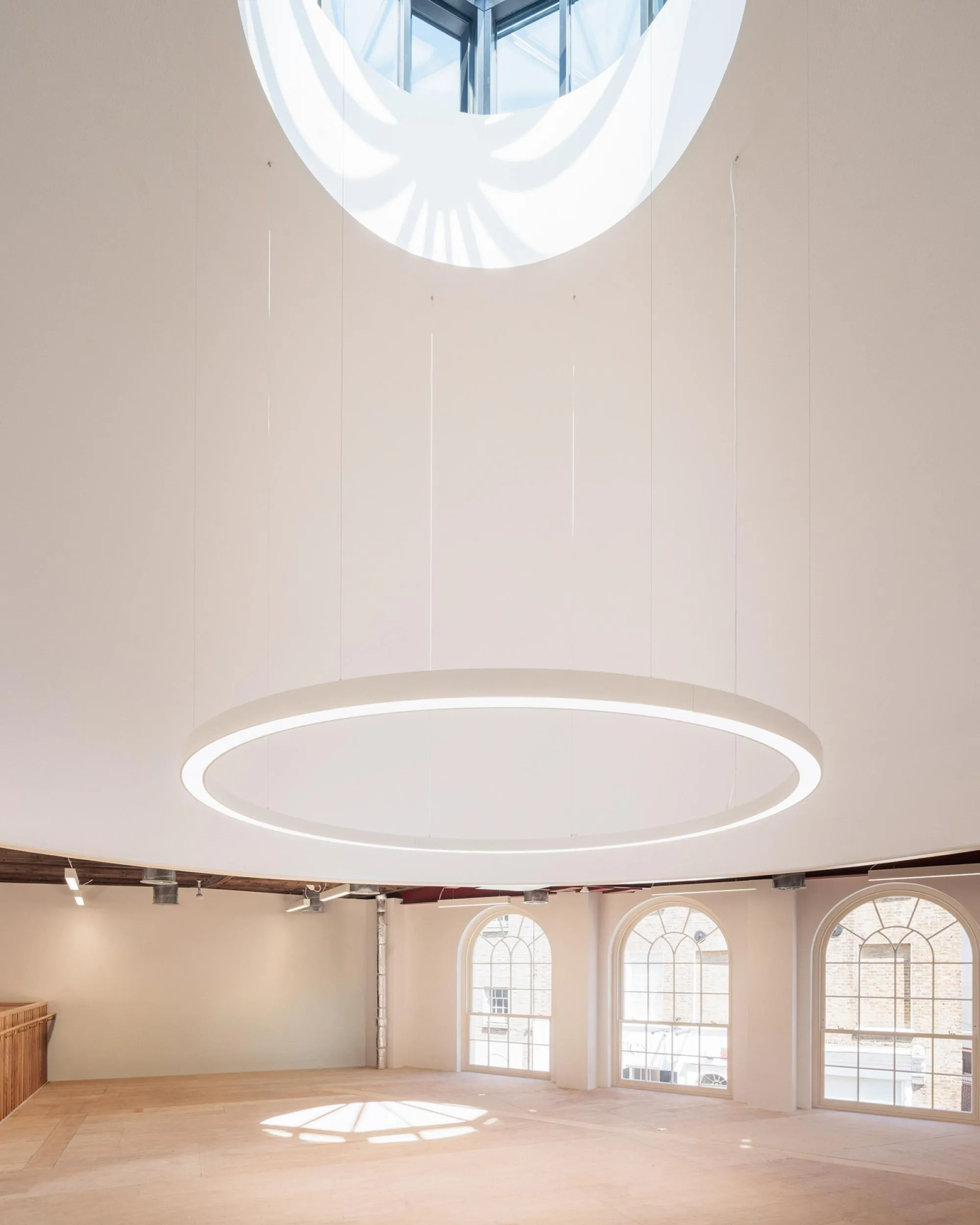 Spacious minimalistic white room with a large circular hanging light and three arched windows allowing natural light inside.
