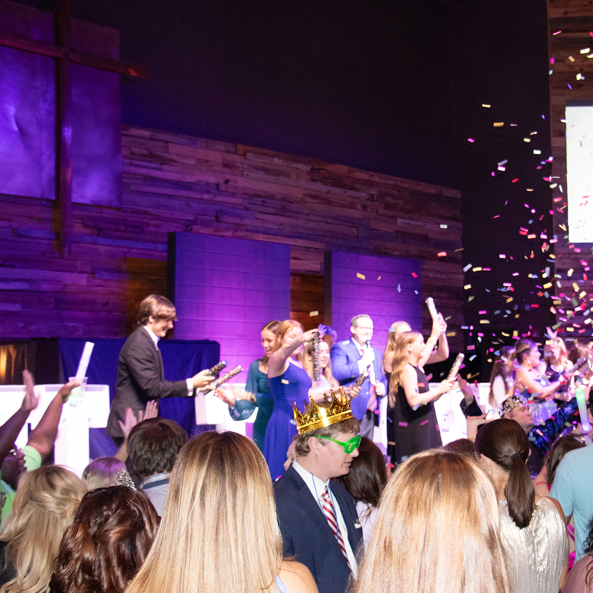 People on stage celebrating with confetti and party poppers in front of a wooden wall with a cross, while a crowd watches.