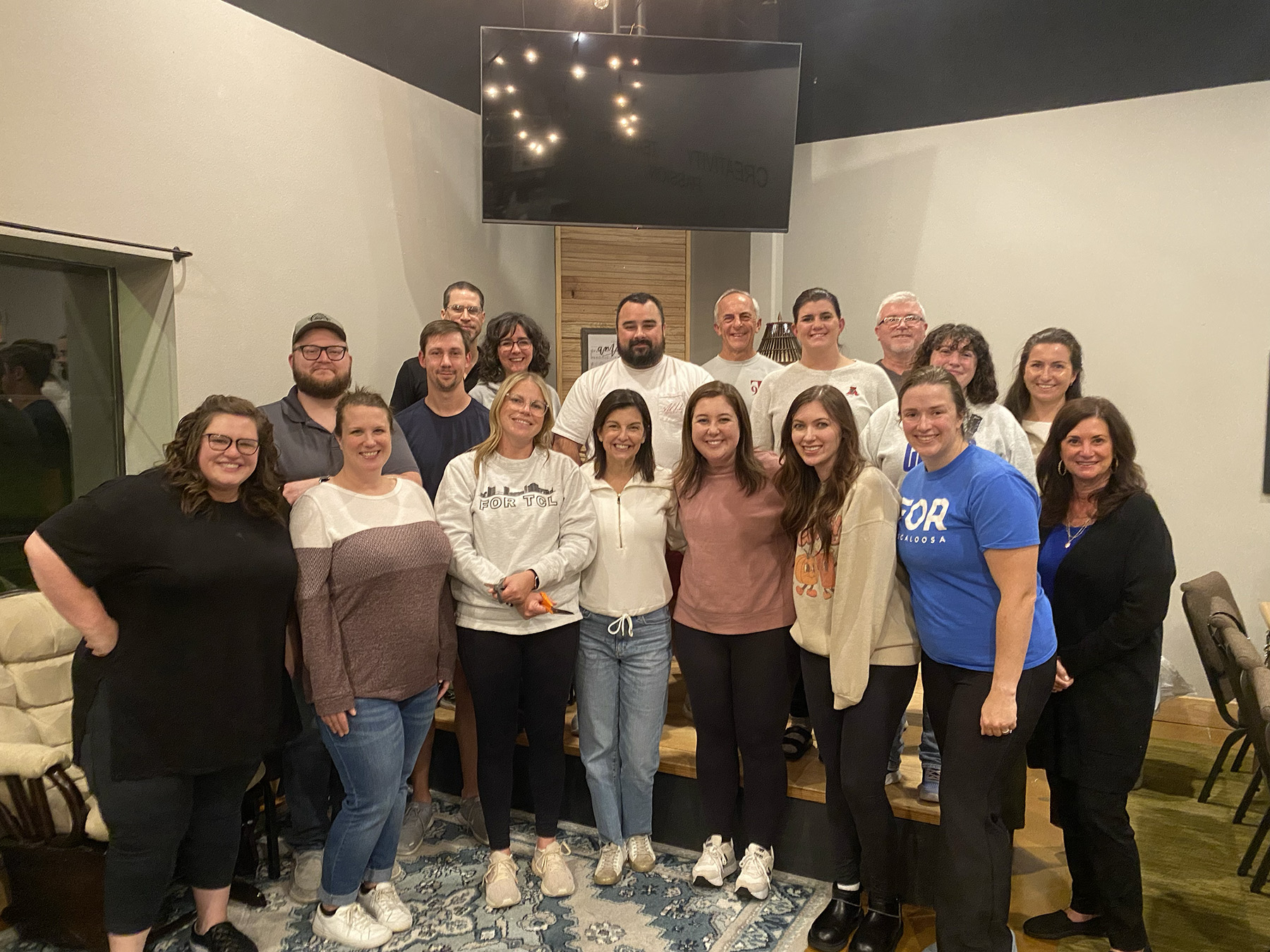 Group of 18 adults smiling and posing indoors in front of a large TV screen mounted on the wall.