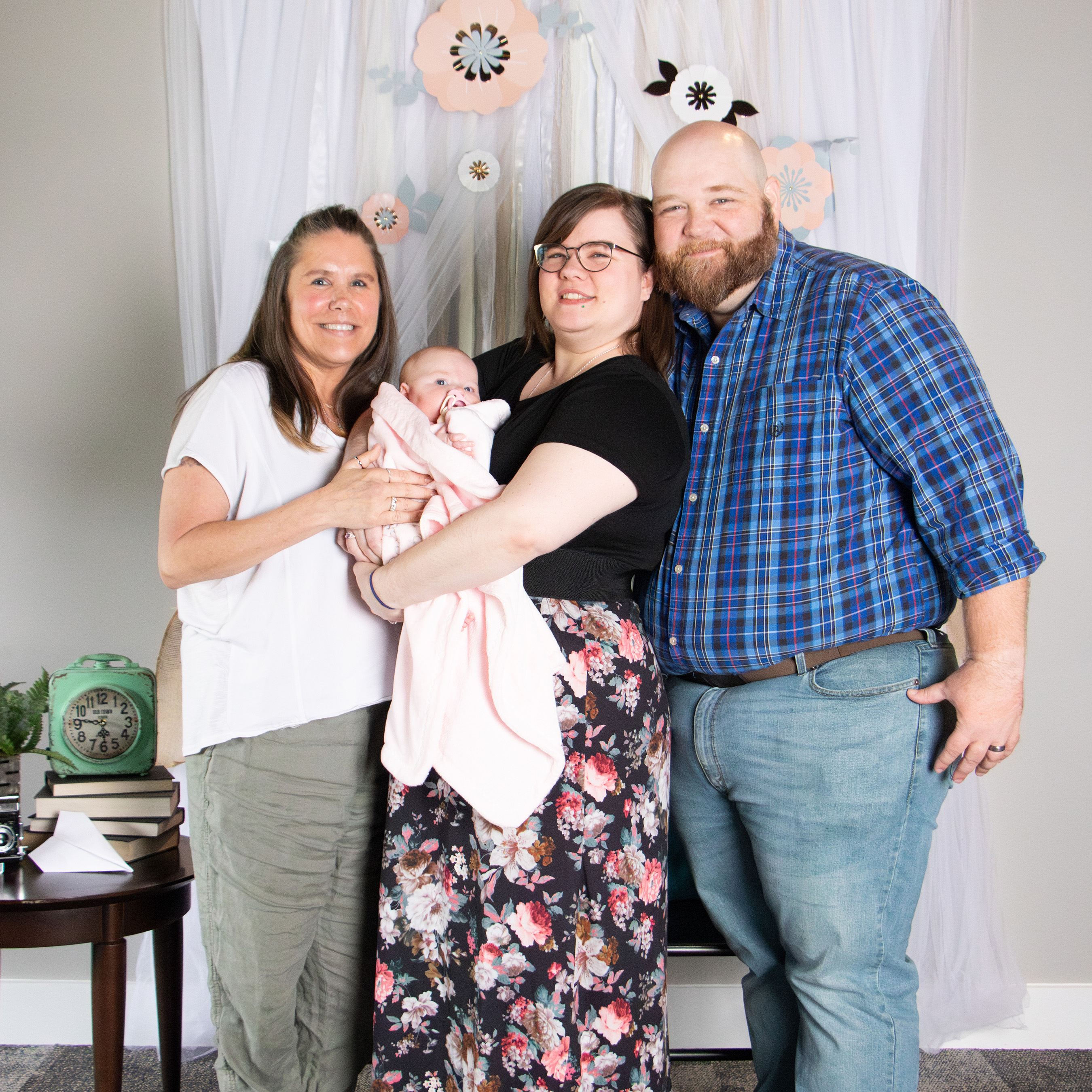 Smiling woman in white blouse, a woman in black holding a baby wrapped in a pink blanket, and a man in a blue plaid shirt standing together in front of a white curtain with flower decorations.
