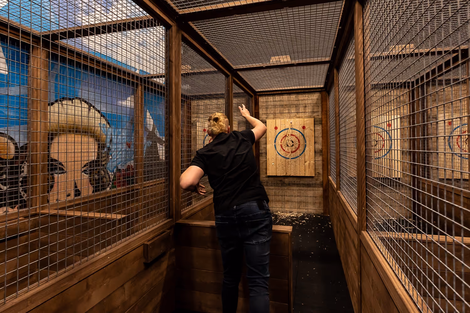 Persoon gooit een bijl naar een houten doelwit met gekleurde cirkels in een omheinde binnenruimte.