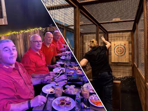 Vier mannen in rode shirts zitten aan een tafel met gegrild eten, terwijl een man in een aparte ruimte een bijl werpt naar een doelwit van hout.
