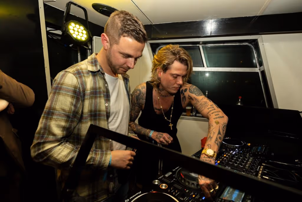 Twee mannen bedienen een DJ-set in een binnenruimte, een met geruite overhemd en de ander met een zwart tanktop en tatoeages.