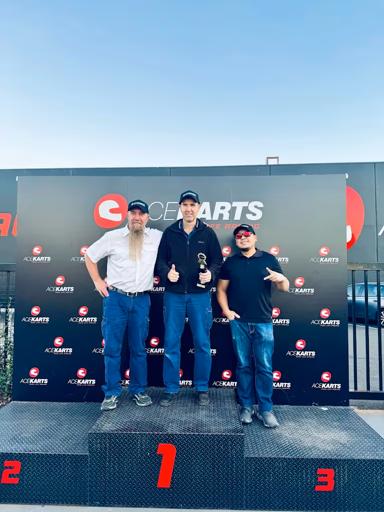 Group of men enjoying food and drinks at an outdoor BBQ area near a go-kart track.