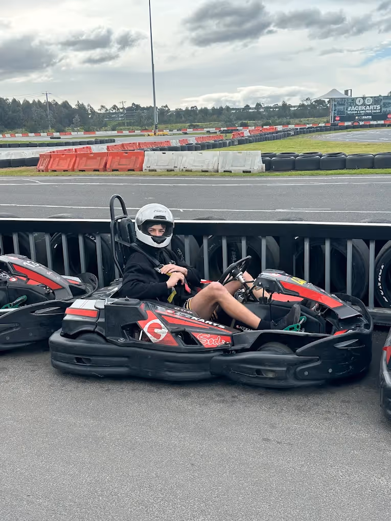 Person wearing a helmet sitting in a red and black Ace Karts go-kart parked beside the track barriers on a cloudy day.