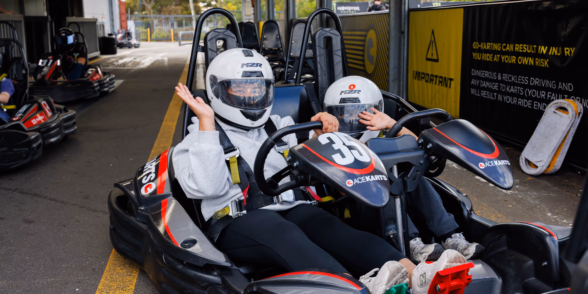 Adult and child wearing helmets seated together in a two-seater Ace Karts go-kart, preparing to drive.