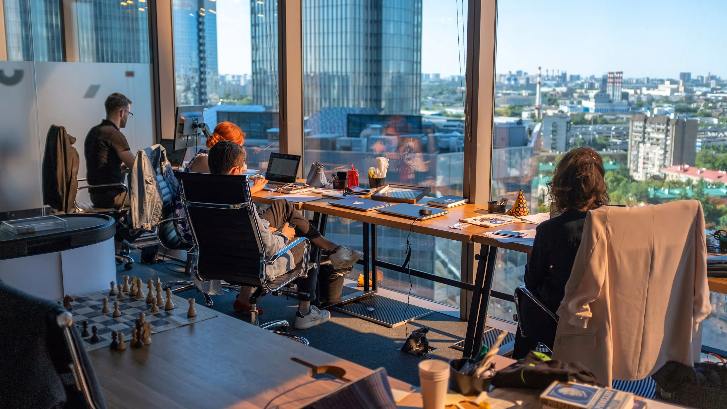 Office space with four people working at desks facing large windows overlooking a cityscape with tall buildings.
