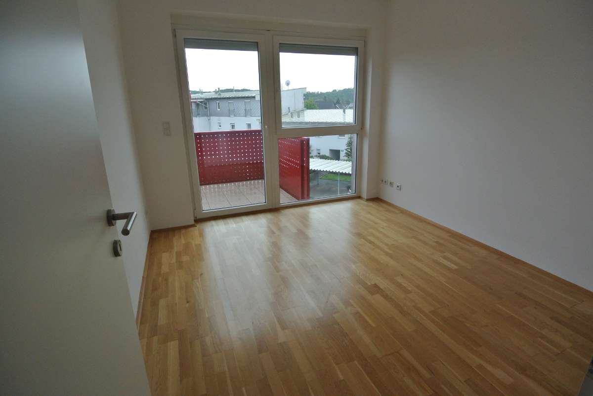 Wohnraum mit großem Fenster und Balkontür, hellem Parkettboden und rotem Balkon-Geländer