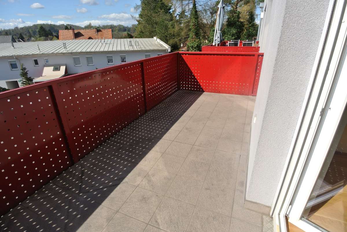 Balkon mit rot gestrichenem Metallgeländer und Ausblick auf Dächer und Bäume