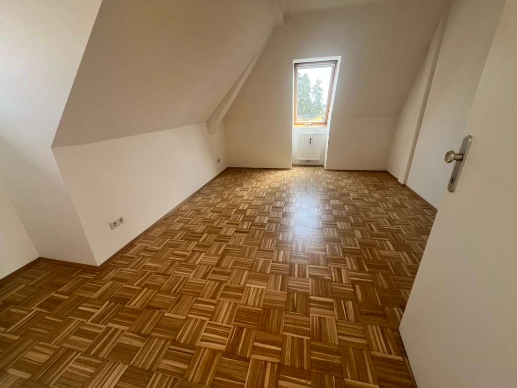 Leeres Zimmer mit Dachschräge, Würfelmuster-Parkett und Fenster mit Heizkörper darunter