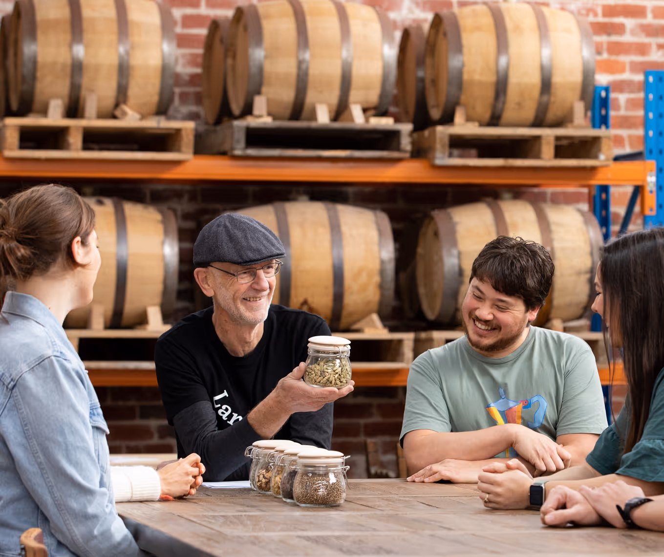 Four people sitting at a wooden table in a brewery with barrels behind, one man holding up a jar of hops and smiling.