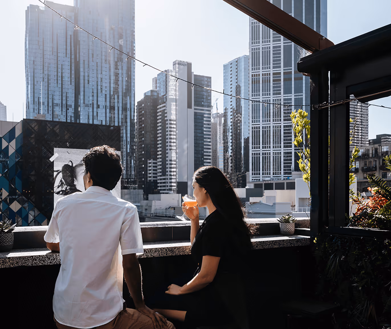 A man and a woman sitting on a rooftop terrace overlooking a city skyline, with the woman drinking from a glass.