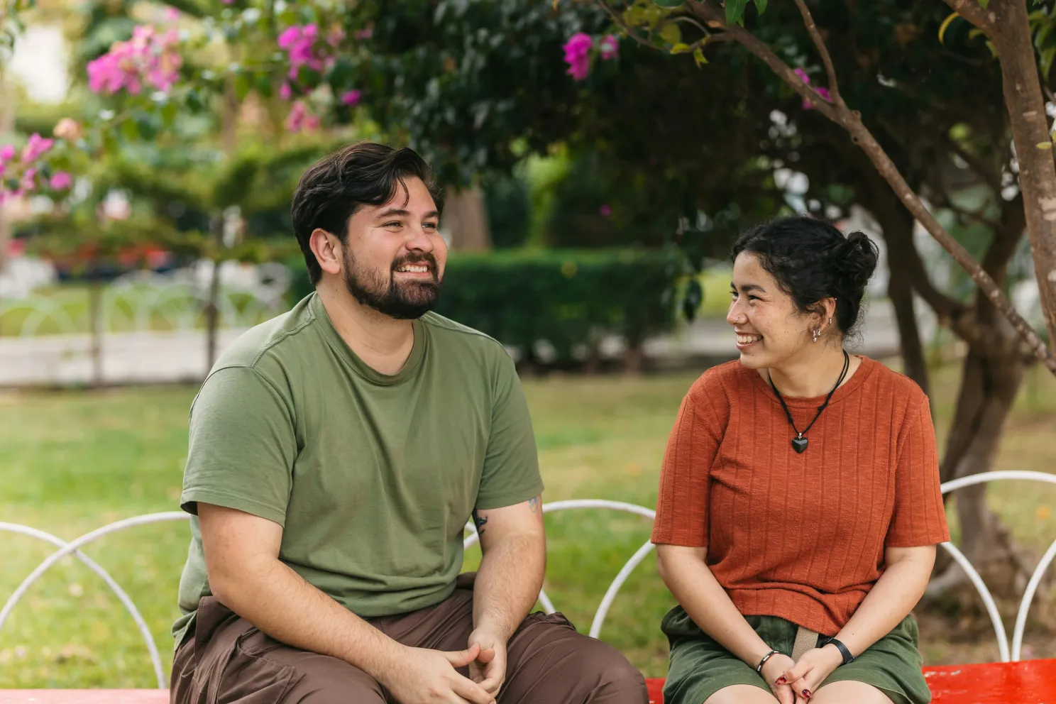 Smiling man in green shirt and woman in orange top sitting and chatting on a red bench in a park.
