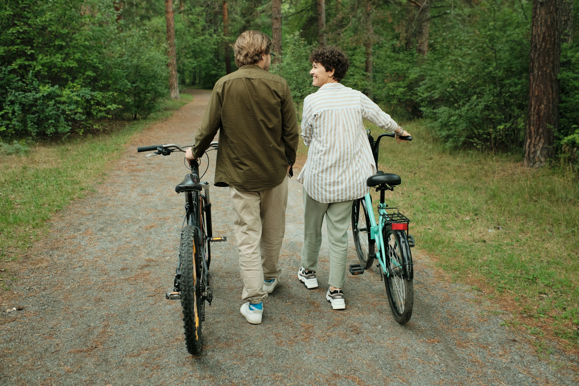 Rear view of a man and woman walking bicycles on a forest path, smiling and talking.
