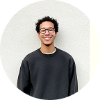Smiling young man wearing glasses and a dark sweater standing against a light wall.