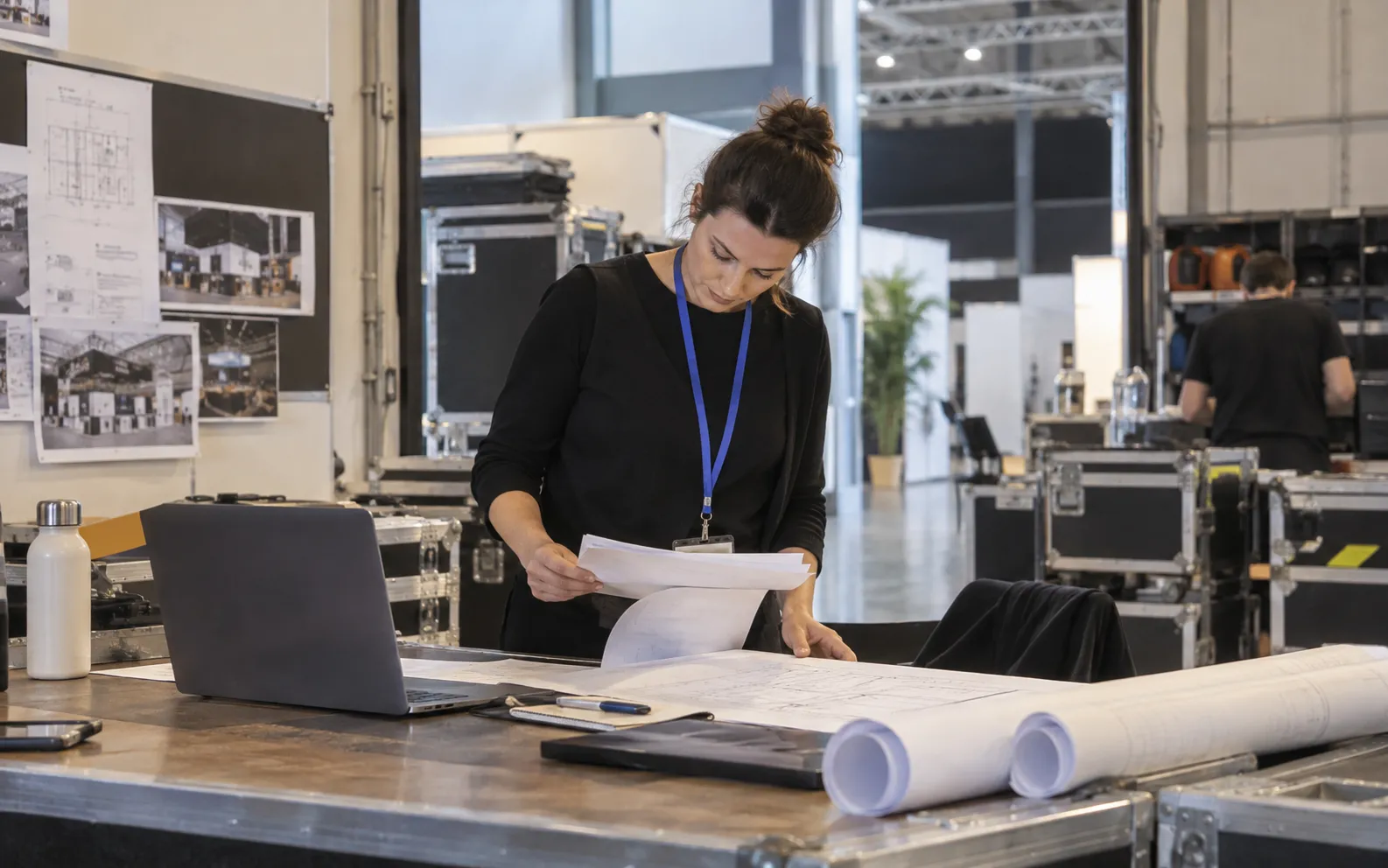 Generated scenario: An exhibition services account director studies a late project brief beside rolled floorplans and equipment flight cases in a venue service office