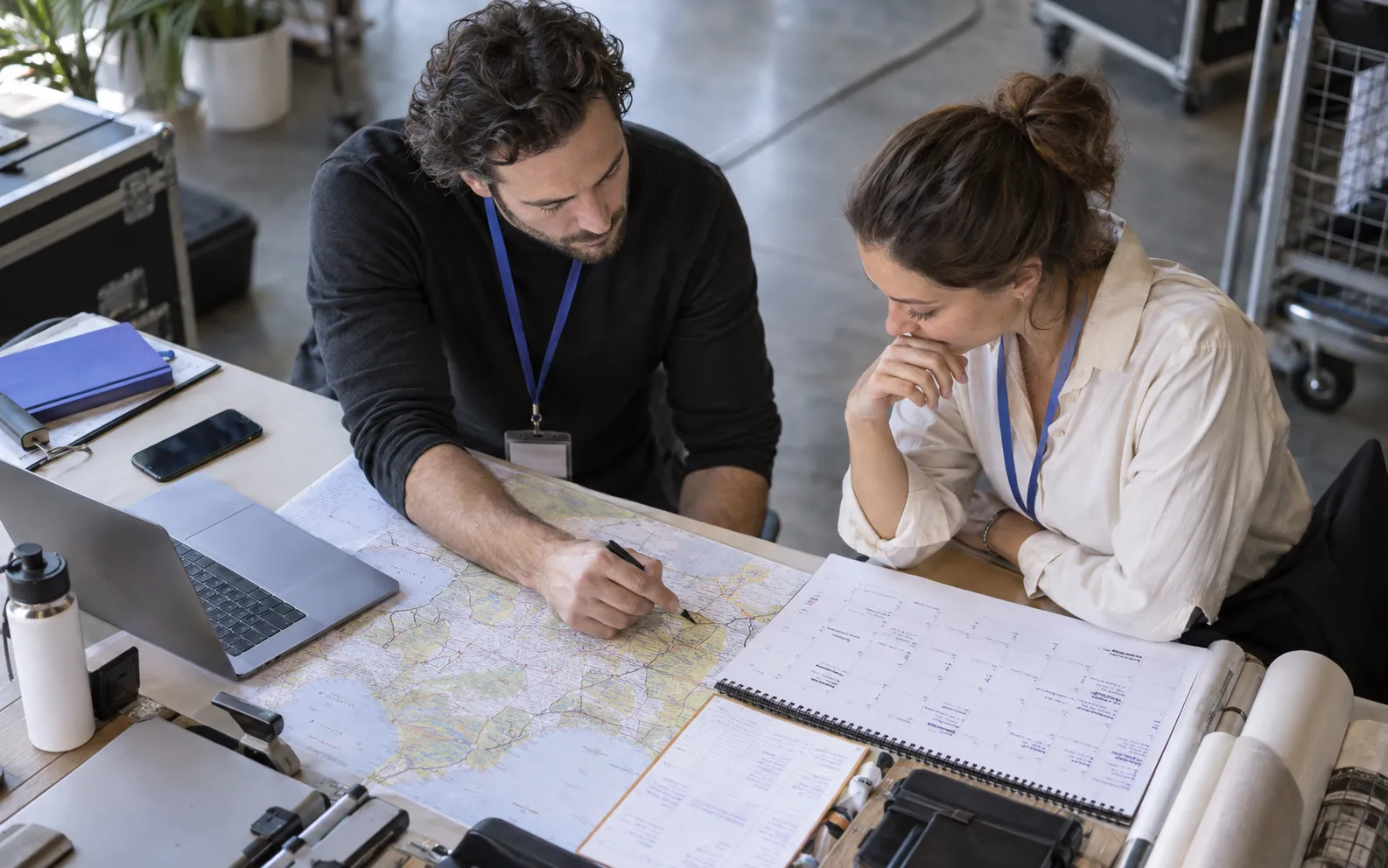Generated scenario: Two agency colleagues check a large physical regional map, a venue calendar and available crew notes on a worktable, considering which exhibitions they can realistically serve
