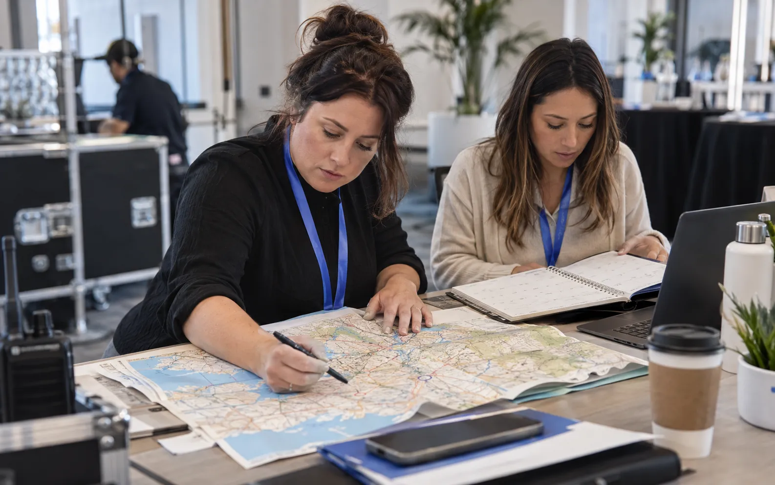 Generated scenario: An event services planner marks two relevant venues on a physical regional map while a colleague checks an open printed calendar, at a venue operations desk