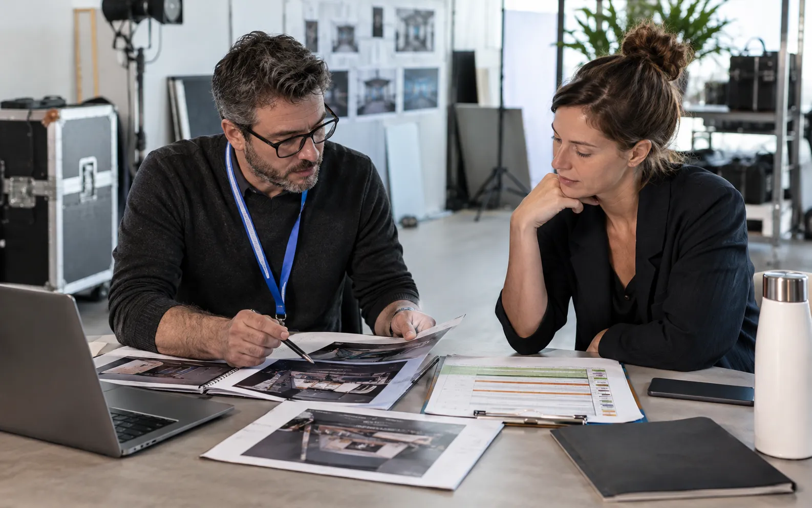 Generated scenario: A producer and account director review three prospective exhibitor folders alongside an equipment availability schedule in a studio meeting area