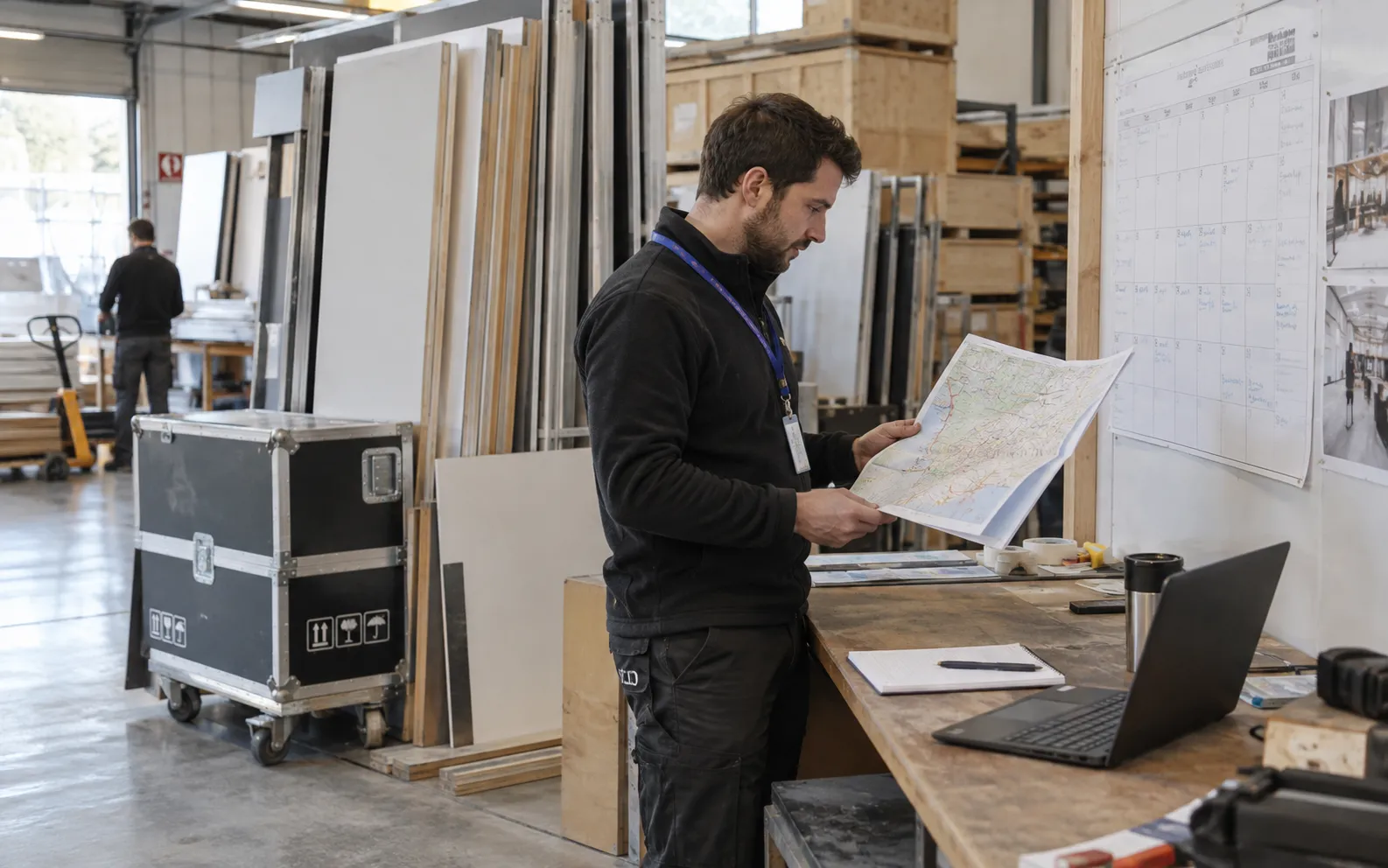 Generated scenario: A fabrication planner checks a wall calendar and a folded delivery-region map beside stacked stand panels and packed transport crates in a clean workshop