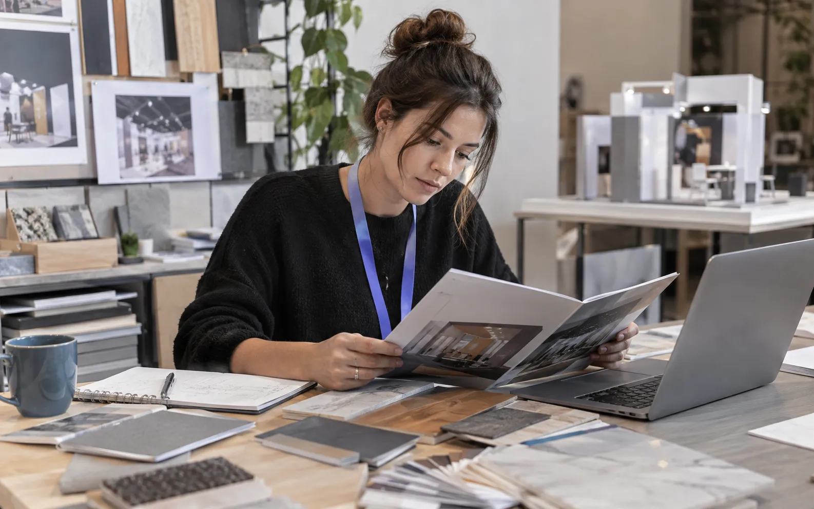 Generated scenario: An exhibit design researcher studies a company product brochure next to material swatches and a laptop in a small practical studio