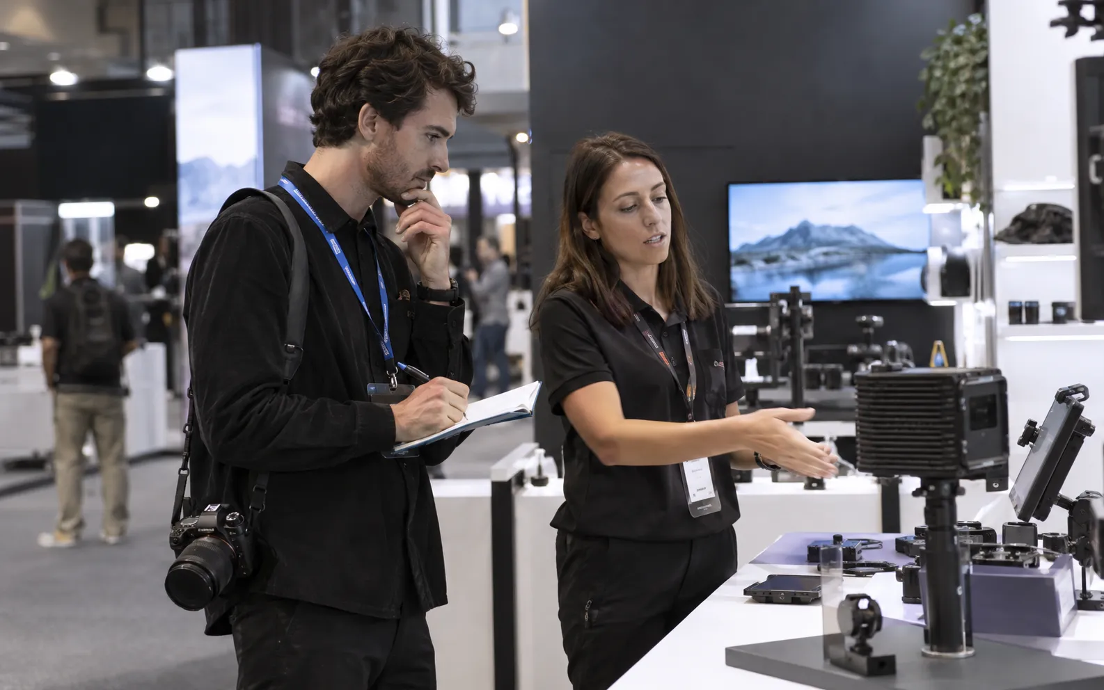 Generated scenario: A film director researches a technical product demonstration in an exhibitor stand while taking unobtrusive notes, a compact camera carried at the side, not filming without permission