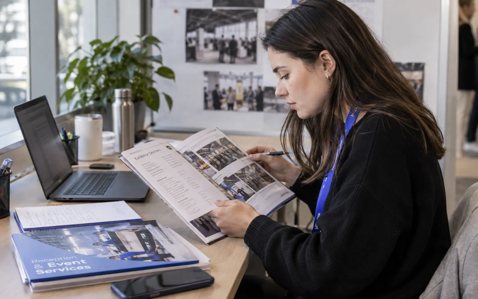 Generated scenario: An event staffing researcher reviews exhibitor sectors in a printed trade-show catalogue beside a reception-service portfolio at a practical agency desk