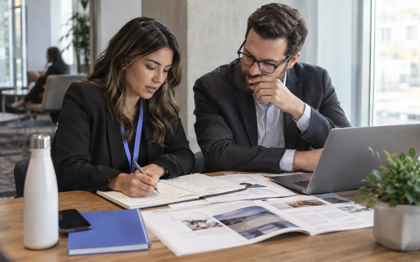 Generated scenario: A demand generation strategist defines a campaign audience on paper with a sales colleague, a printed industry event guide and company research spread on a planning desk