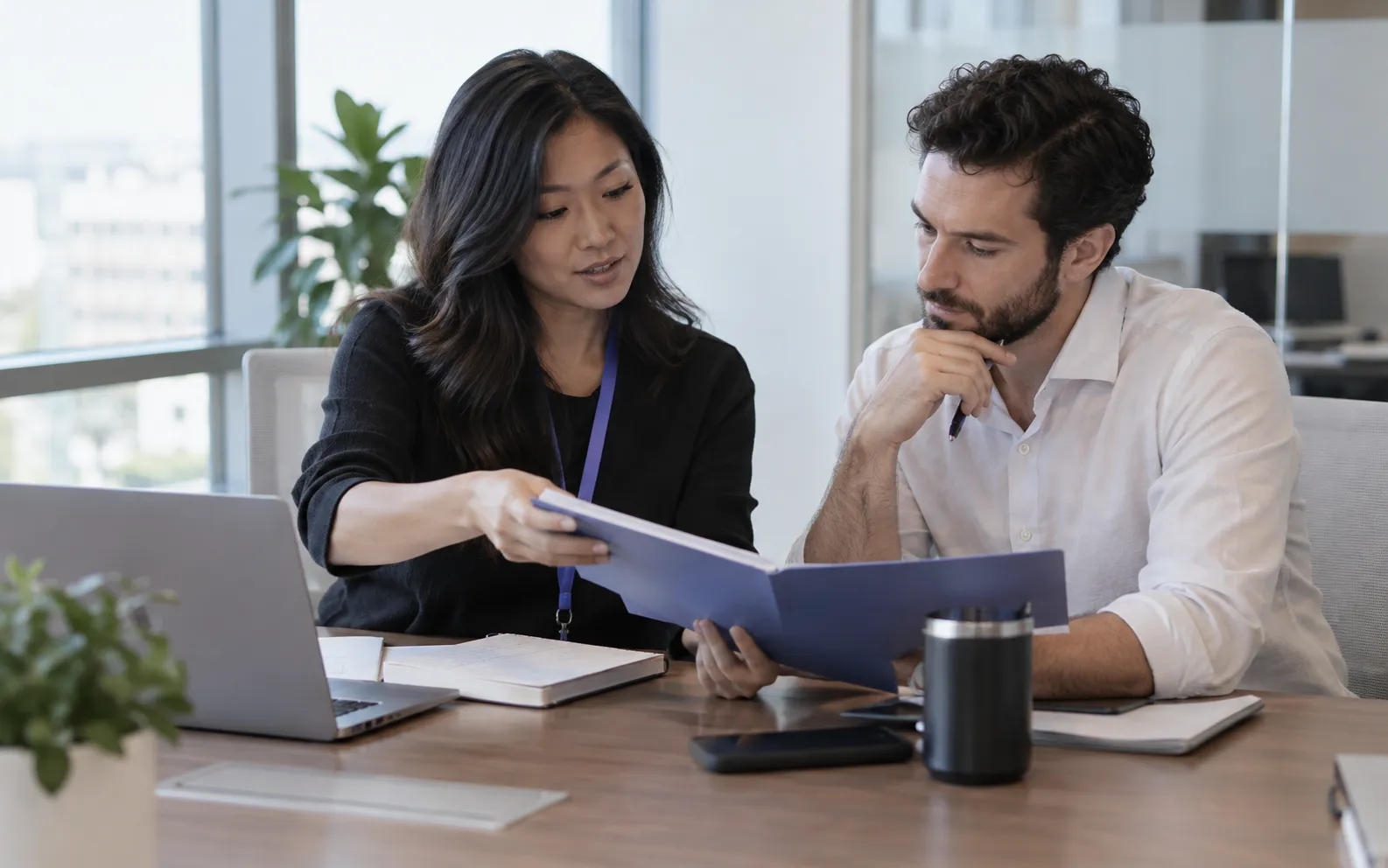 Generated scenario: A demand generation lead passes a researched account brief to a sales colleague at an office meeting table, both reviewing the same folder and next-step notes