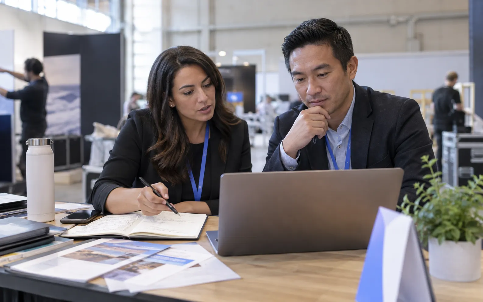 Generated scenario: An event marketer coordinates a pre-show account handoff on a laptop with a sales colleague, using paper invitation notes and their existing planning materials