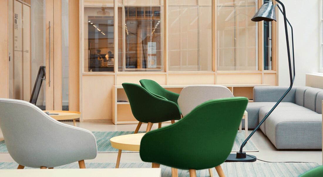 Modern office lounge area with green and gray chairs, wooden tables, a gray sofa, and a tall black floor lamp.