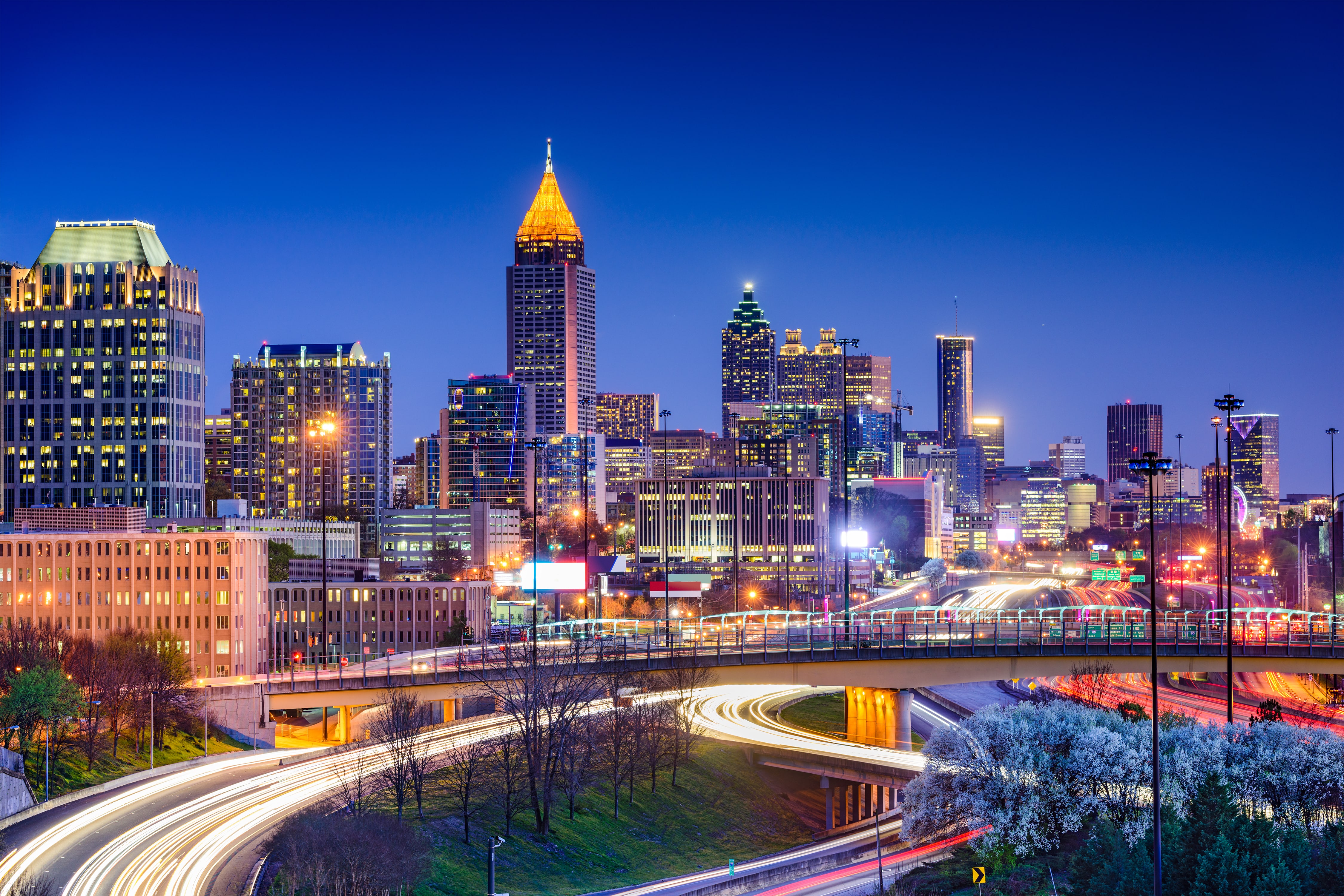 Night view of Atlanta skyline with illuminated skyscrapers and light trails from cars on curving highways.