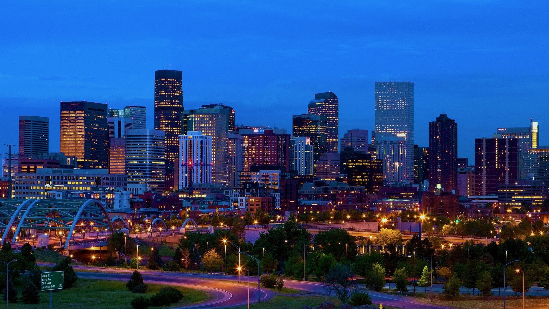 City skyline at dusk with illuminated modern high-rise buildings and a curved road in the foreground.