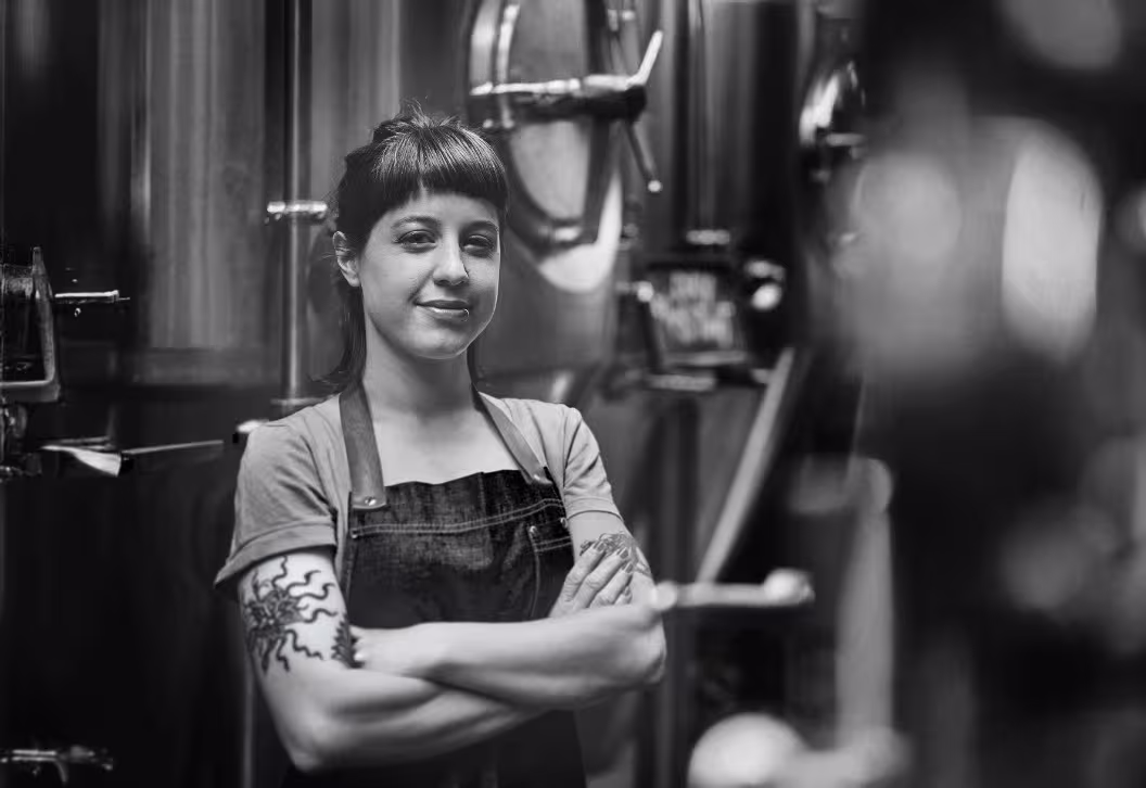 A female craft brewer with tattoos and a denim apron standing confidently in front of brewery tanks, representing independent brewers using the ZANZ canning machine