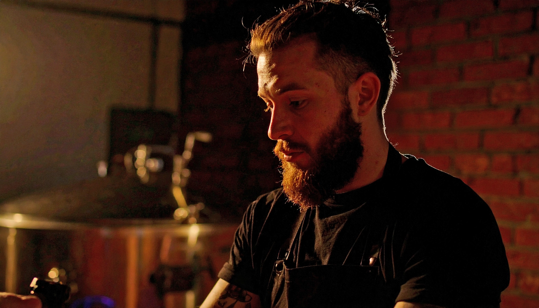 A bearded male brewer in a black apron working alone in a warmly lit brewery with brick walls, representing the elimination of expensive canning expert dependency with the ZANZ Solo