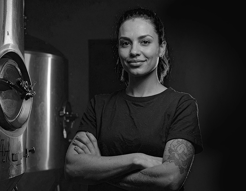 Confident woman with tattooed arm standing with arms crossed near industrial brewing equipment.