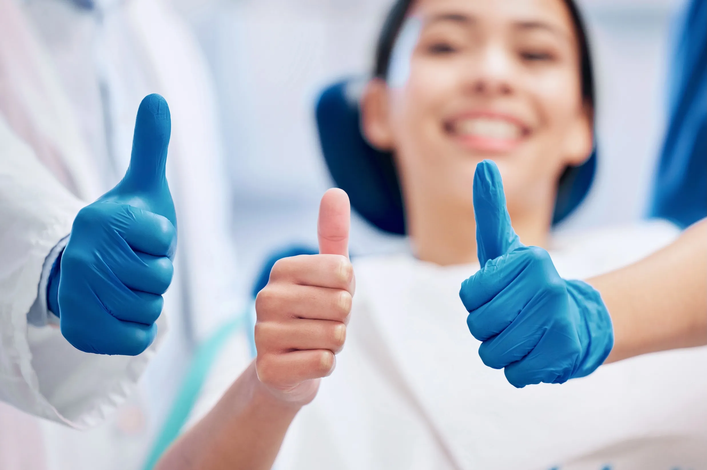 Three thumbs up in close-up, with two wearing blue medical gloves and a smiling person blurred in the background at Azusa Dental Clinic.