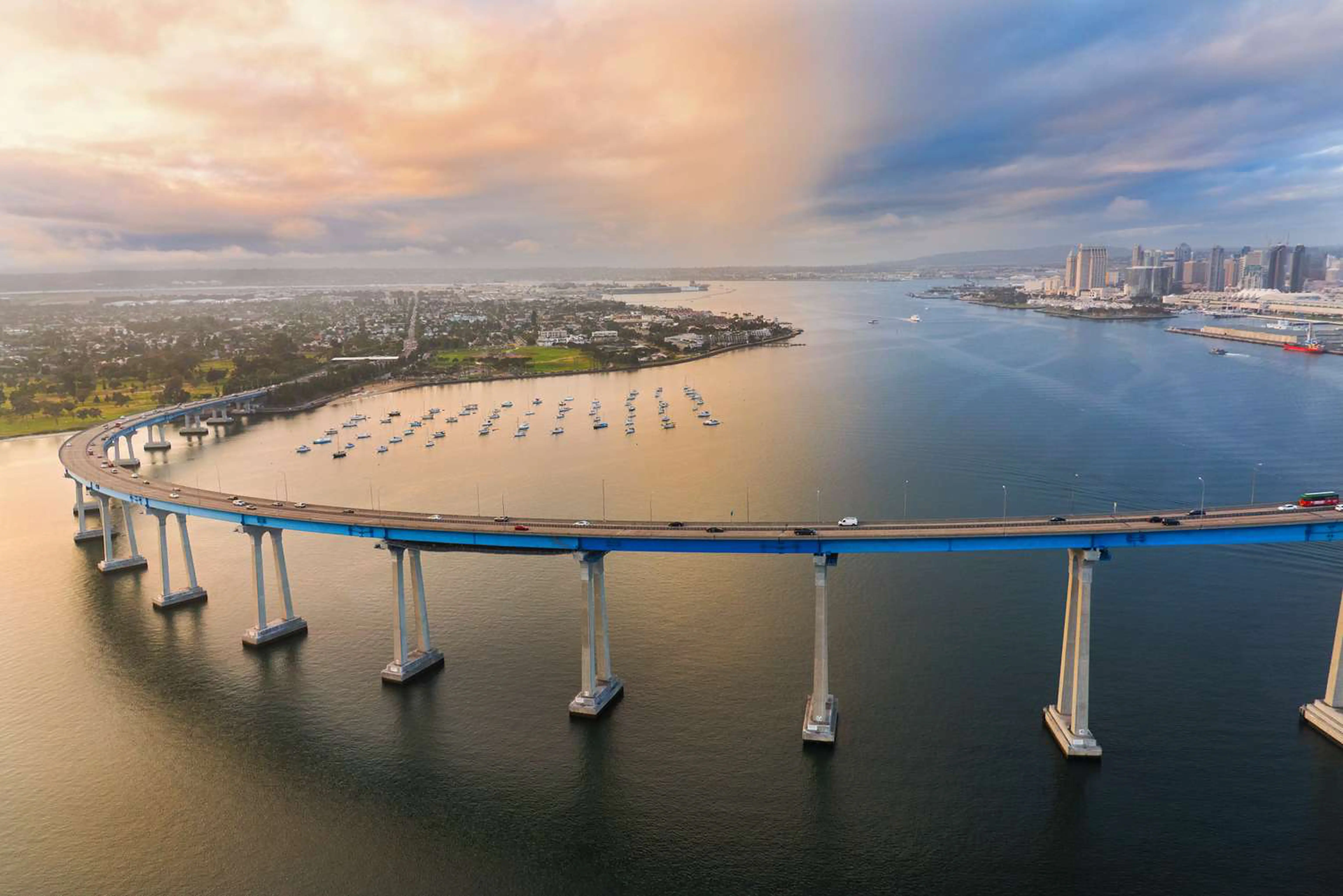 Curved bridge over calm water with boats docked on one side and a city skyline in the background under a partly cloudy sky during sunset.