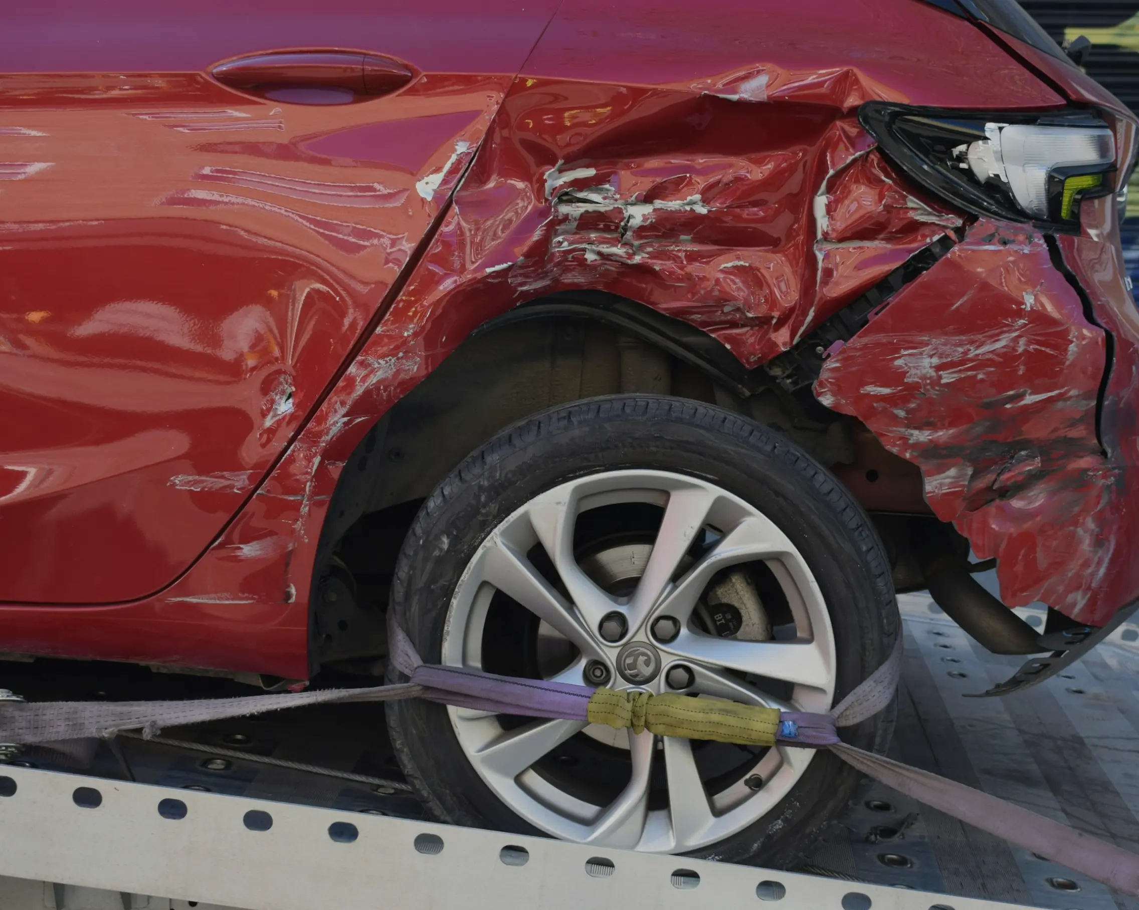 Close-up of a red car's severely damaged front right side with a scratched and crushed fender, secured with straps on a flatbed truck.