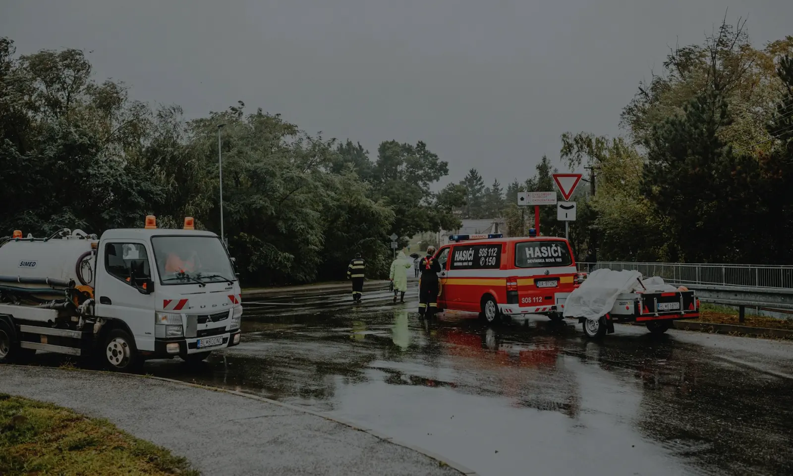 Rain-soaked road with a white tanker truck and a red emergency response van marked Hasiči, with people in rain gear near the vehicles.