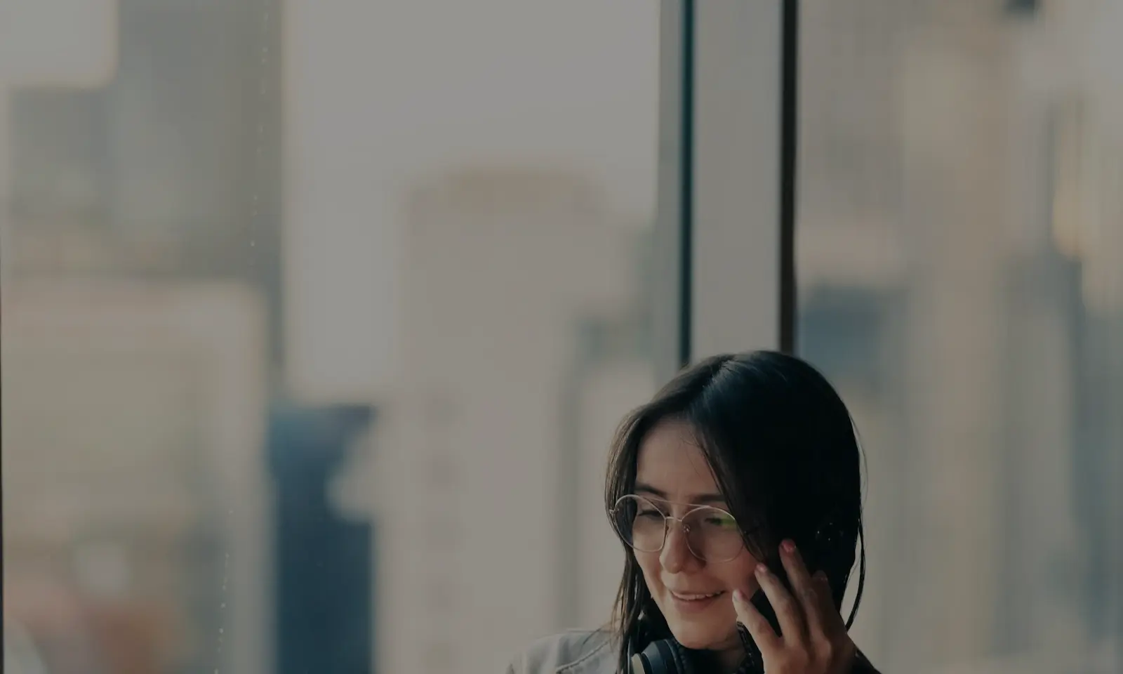 Smiling woman with glasses and headphones talking on a phone by a large window with a blurred cityscape outside.