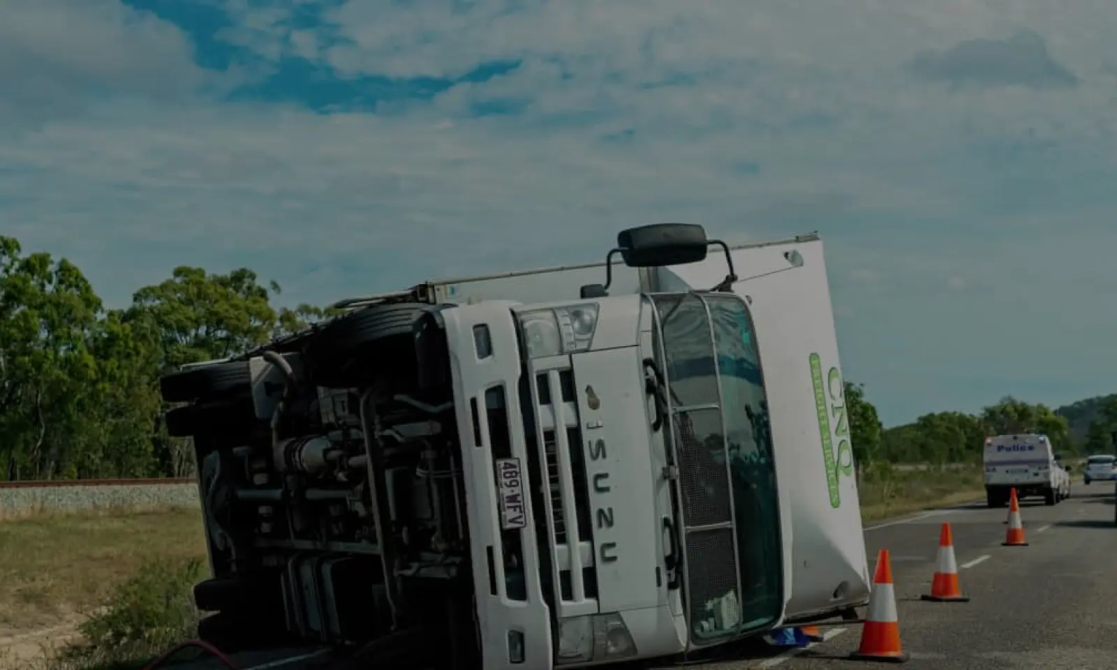 A white Isuzu truck overturned on its side on a rural road with traffic cones and a police vehicle in the background.