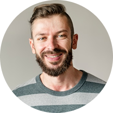 Smiling man with a beard and short hair wearing a gray striped shirt against a plain light background.