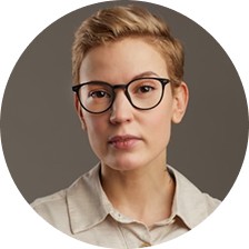 Portrait of a young person with short blonde hair and glasses wearing a beige collared shirt against a neutral background.