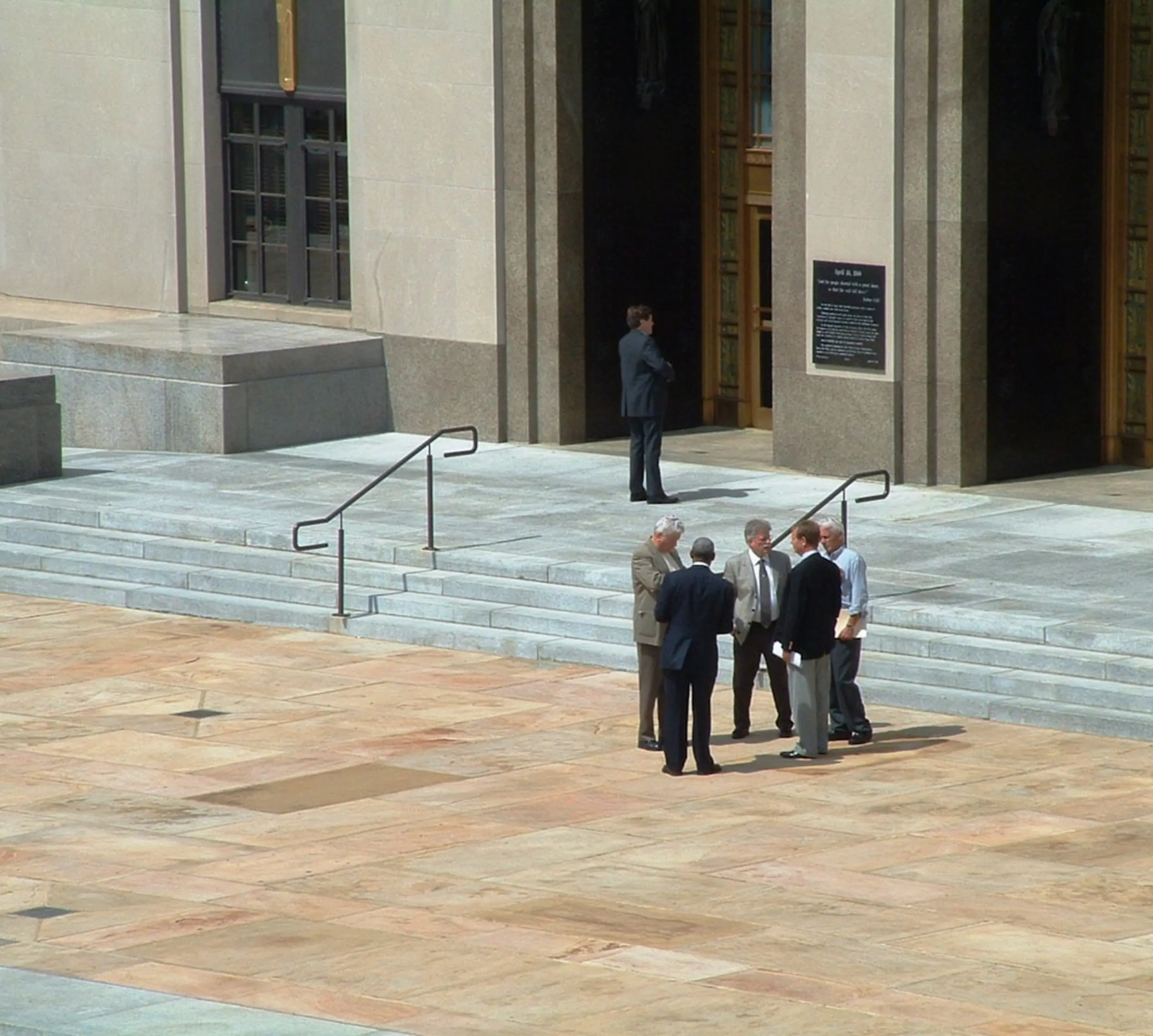Five men in business attire standing and talking on the steps outside a large stone building with tall doors.
