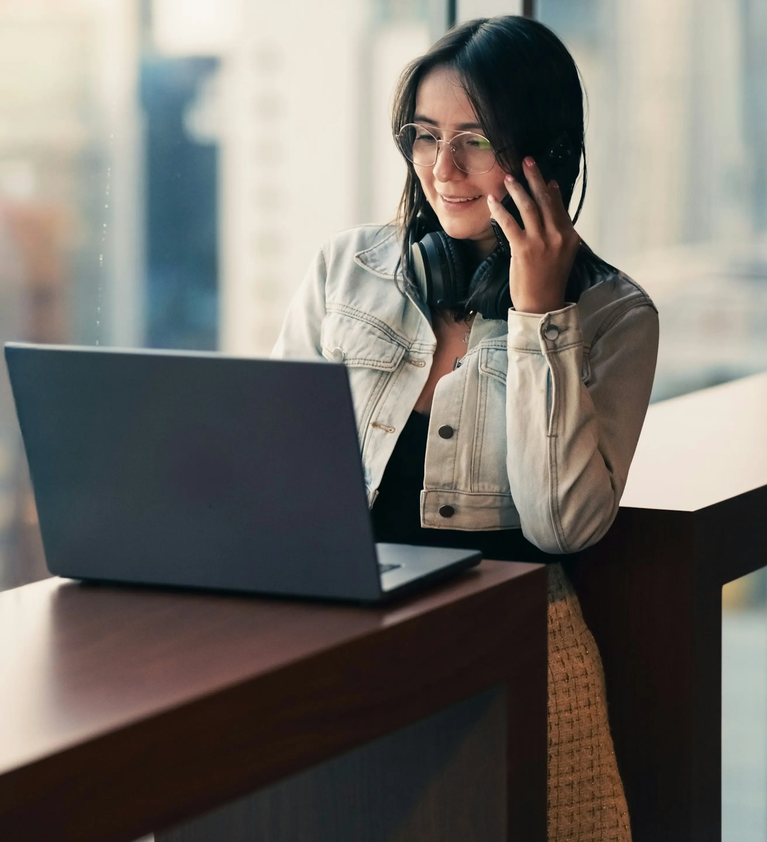 Young woman wearing glasses and a light jacket talks on the phone while using a laptop at a wooden desk.