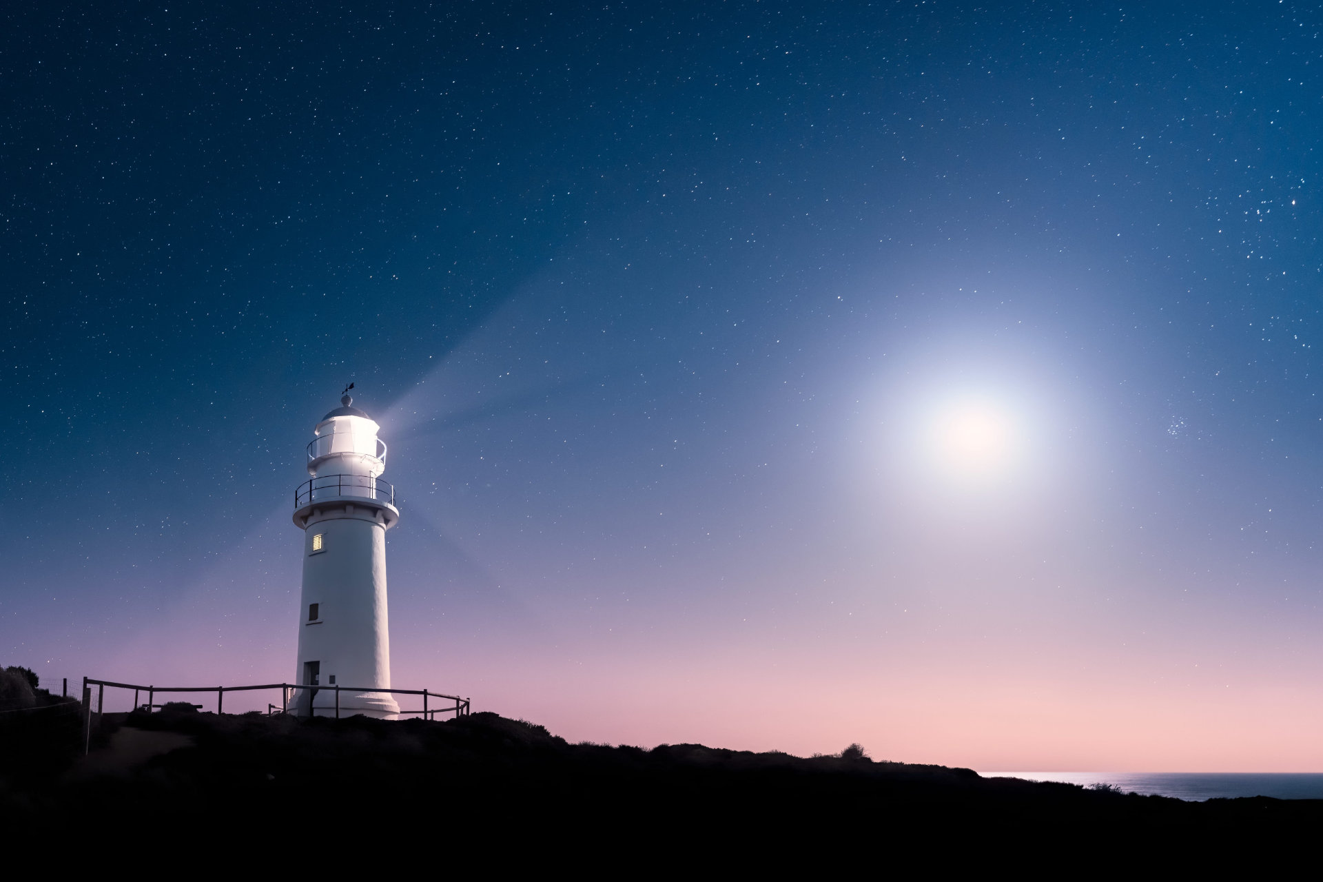 White lighthouse shining its light under a starry night sky with a bright moon over dark silhouette of land and ocean. Yorke Peninsula Corny Point.