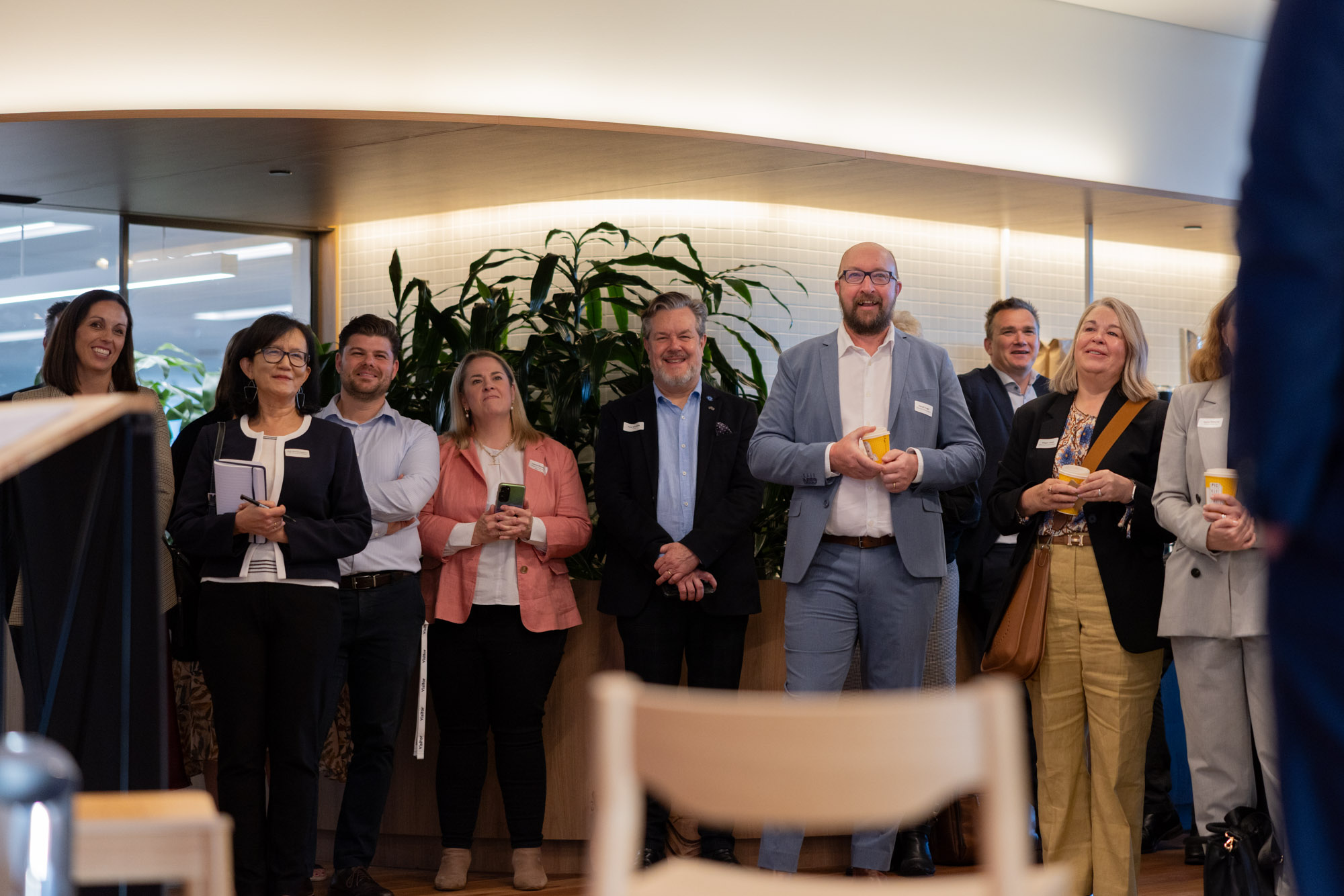 Group of business professionals standing indoors, some holding coffee cups, attentively listening to a speaker.